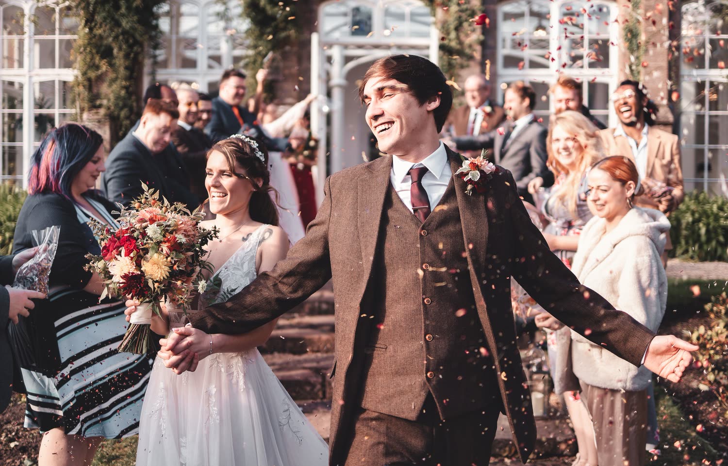 Bride and groom laughing through a confetti throw outside a Victorian glasshouse, the groom in brown tweed and the bride carrying an autumn bouquet, wedding photography